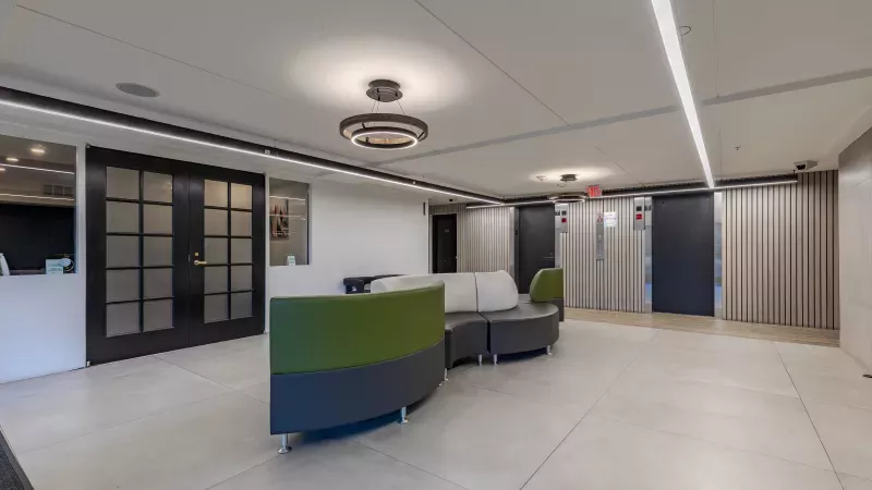 Modern office lobby with a curved green and gray sofa, dark doors, and two silver elevators.