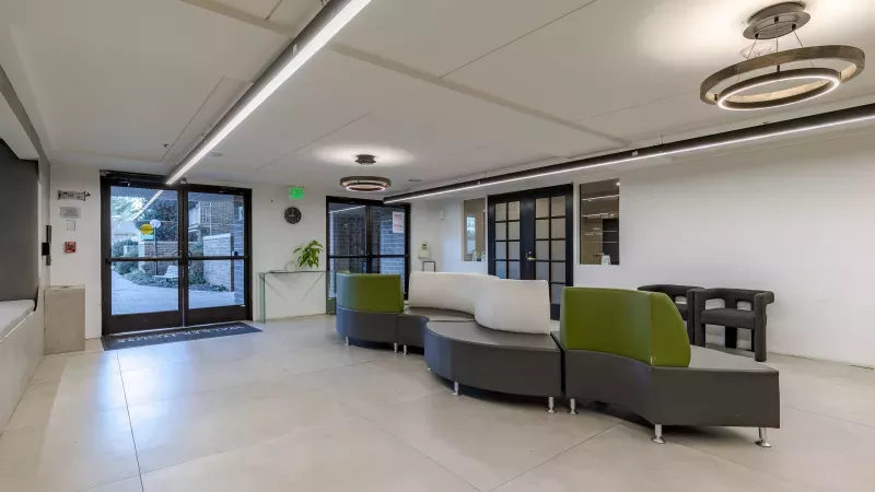 Modern lobby with light-colored floors, curved gray and green seating, and circular ceiling lights near double glass doors.