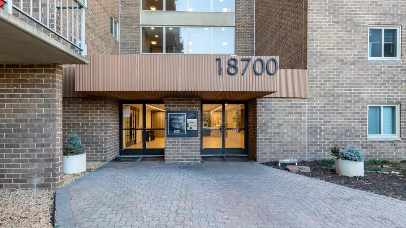 Main entrance of a brick apartment building featuring double glass doors, a wooden awning, and the address '18700'.