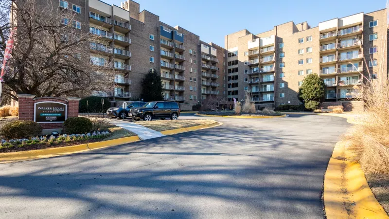 Large brown apartment building with multiple balconies, a circular driveway, parked cars, and winter trees.