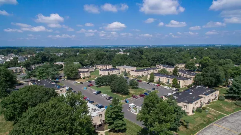 Aerial photograph of residential buildings with light facades, dark roofs, cars in parking lots, and many green trees.