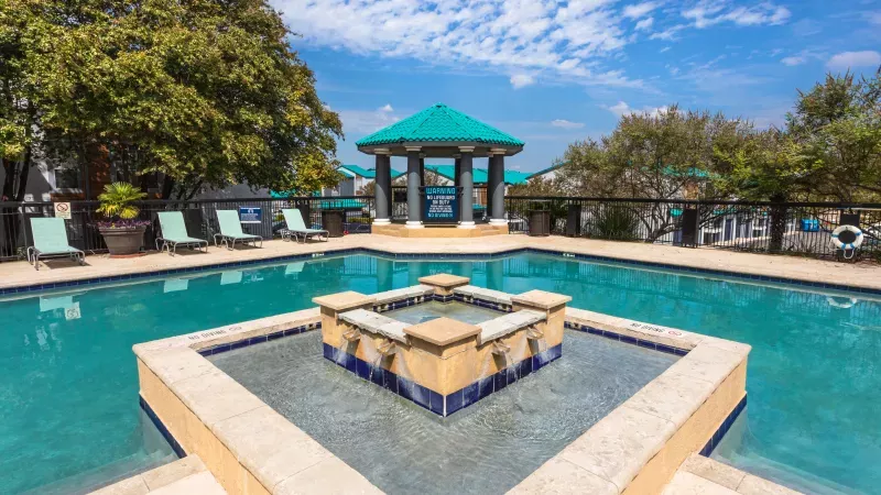 Outdoor swimming pool with a stone fountain, lounge chairs, and a turquoise-roofed gazebo under a blue sky.