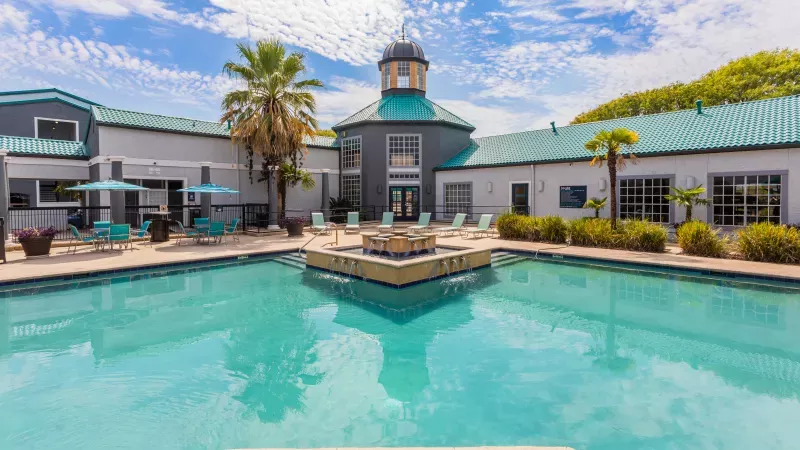 Vibrant outdoor swimming pool with a central dome building, surrounded by teal-roofed apartments, palm trees, and lounge areas.