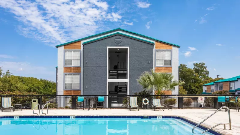 Sunny day view of a modern apartment building with gray siding and teal trim, overlooking a bright blue swimming pool.