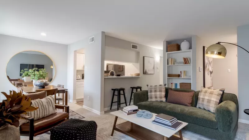Modern living room with a green sofa, coffee table, bar, and a dining area visible through a large round mirror.