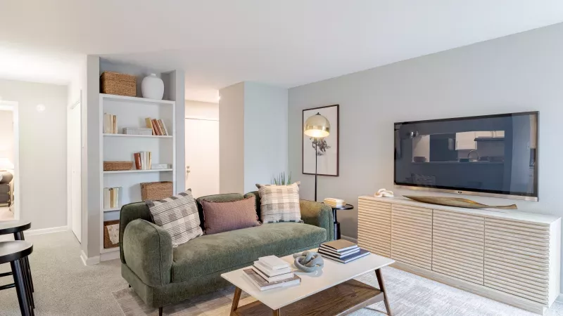 Modern living room with a green sofa, coffee table, TV on a light wood stand, and a white bookshelf.