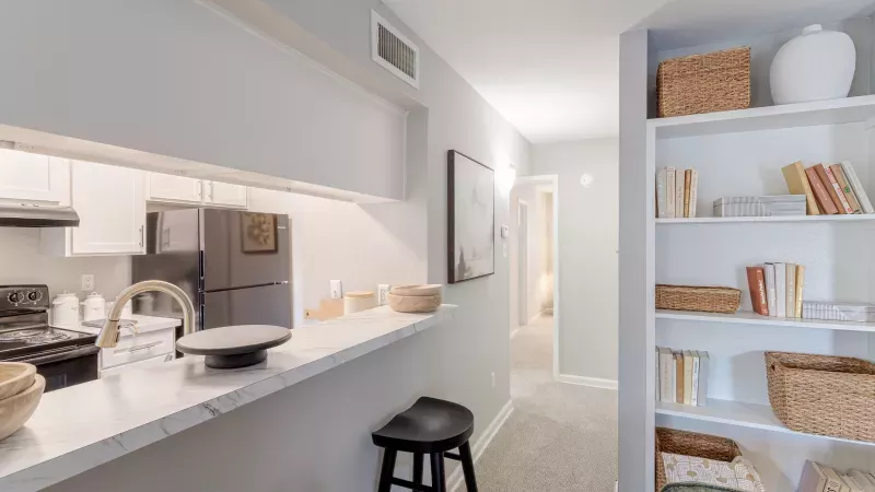 Modern apartment kitchen with white cabinets, black appliances, and breakfast bar, beside a white bookshelf.