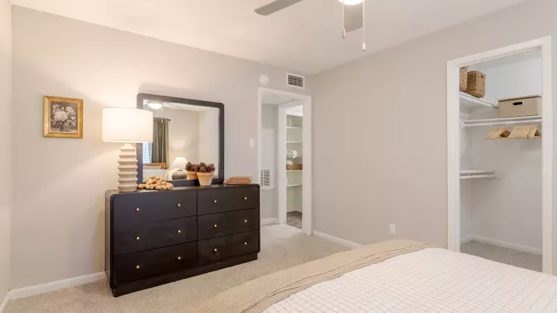 A neutral-toned bedroom features a dark dresser with a mirror, a bedside lamp, and a bed with white and beige bedding.