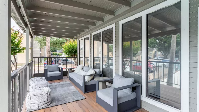 Modern outdoor porch with grey sofa, armchairs, and ottomans on a wooden deck, overlooking a tree-lined street.