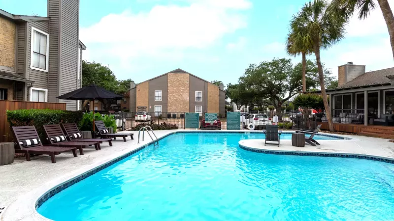 Vibrant blue swimming pool surrounded by dark lounge chairs, apartment buildings, and palm trees under a partly cloudy sky.