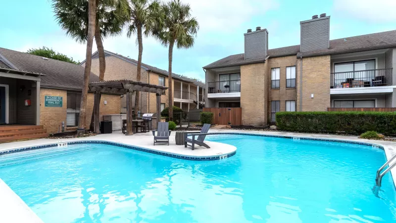 Outdoor swimming pool with light blue water, surrounded by apartment buildings, palm trees, and a shaded seating area.