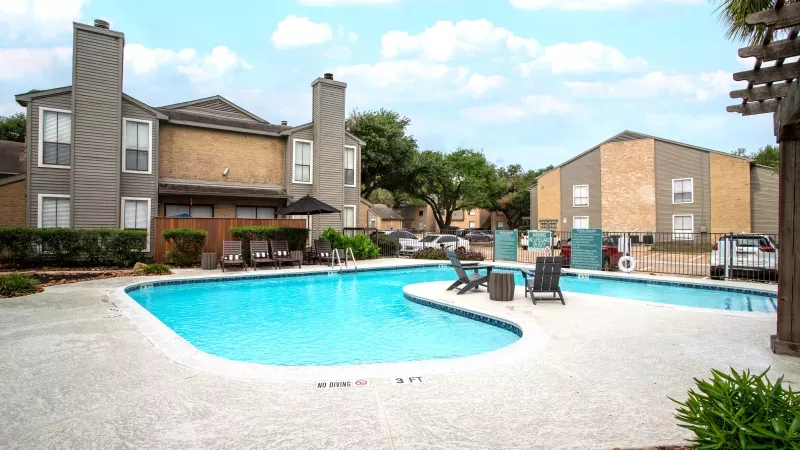 Apartment complex pool with blue water, lounge chairs, and tan buildings under a partly cloudy sky.