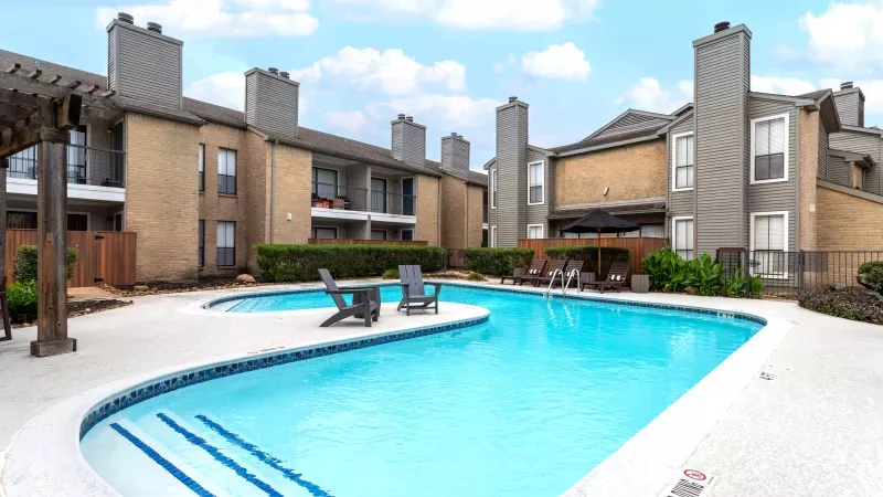 Sunny outdoor swimming pool with blue water, lounge chairs, and multi-story apartment buildings.