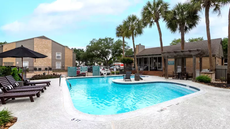 Photograph of an outdoor swimming pool surrounded by lounge chairs, palm trees, apartment buildings, and a clubhouse.