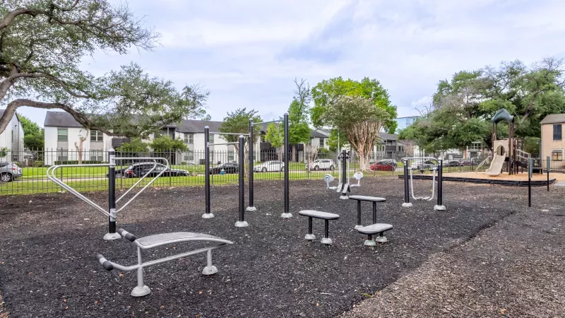Outdoor fitness park with various exercise equipment on dark rubber surfacing, surrounded by trees and apartment buildings.