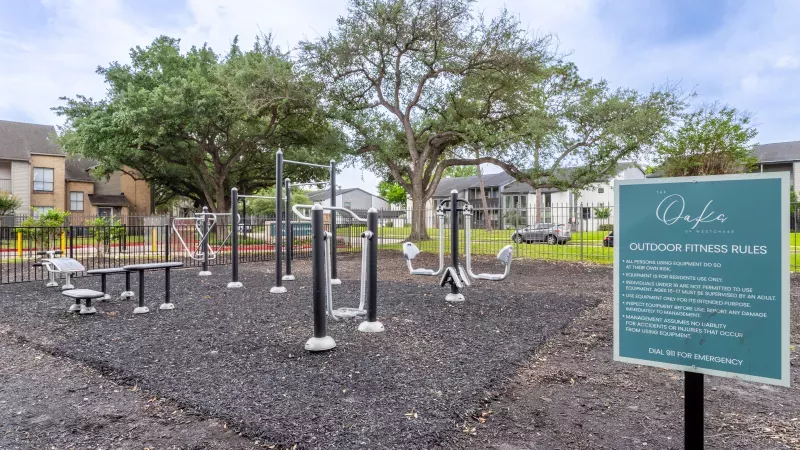 Outdoor fitness area with silver and black exercise equipment on black ground, with trees and residential buildings in the background.