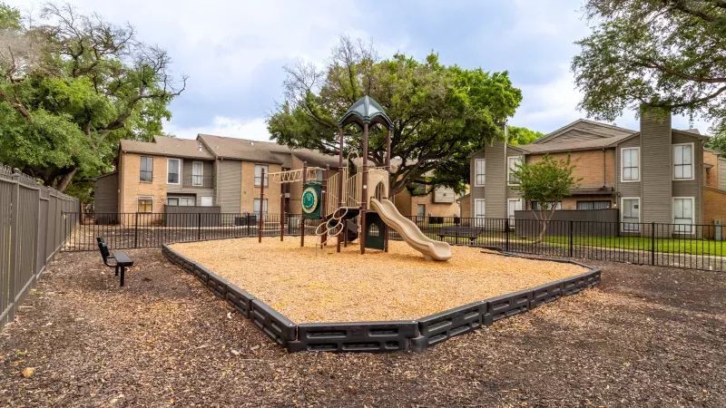 Playground with a large climbing structure and slide on wood chips, backed by trees and apartment buildings under a cloudy sky.