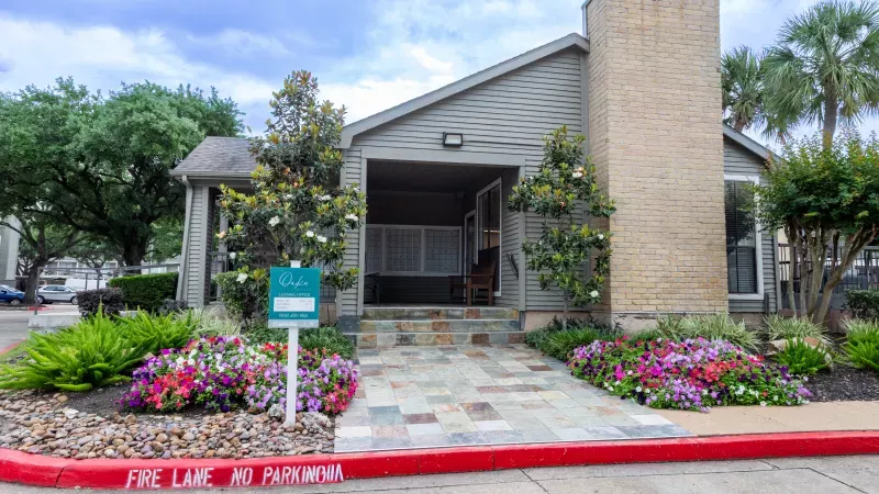 Grey building with a brick chimney, stone path, and colorful flower beds. A red fire lane curb is in the foreground.