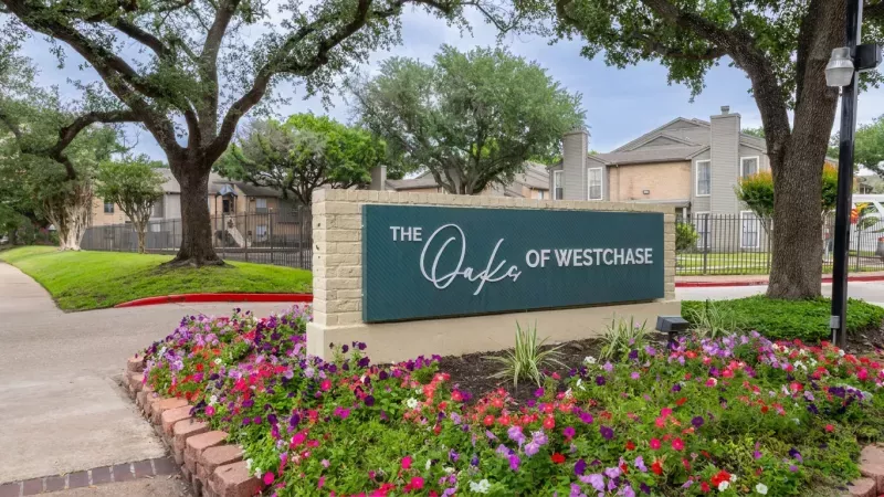Green sign reading "The Oaks of Westchase" at an apartment complex entrance, bordered by colorful flowers and trees.