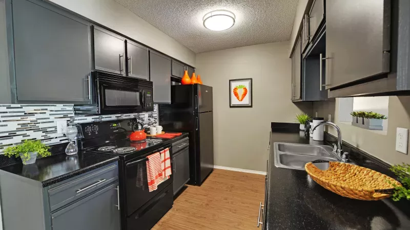 Modern kitchen with dark gray cabinets, black appliances, and countertops. Features a patterned backsplash, wood-look floor, and orange decor.