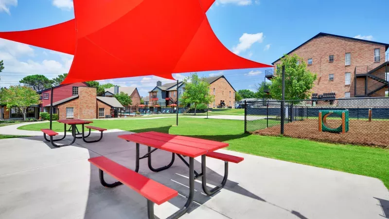 Red picnic tables under red shade sails on a patio, with grass, a dog park, and apartment buildings.