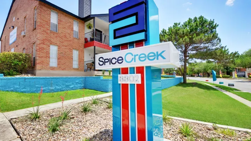 Vertical blue and red Spice Creek apartment sign with its logo and "5102," a brick building and green lawn behind.
