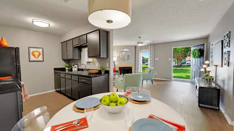 Bright open-concept apartment with a dining table, modern dark kitchen, and living room opening to a green outdoor lawn.