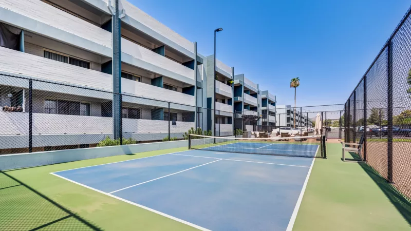 Blue and green tennis court with a net, flanked by a fence, in front of a modern apartment building under a clear sky.