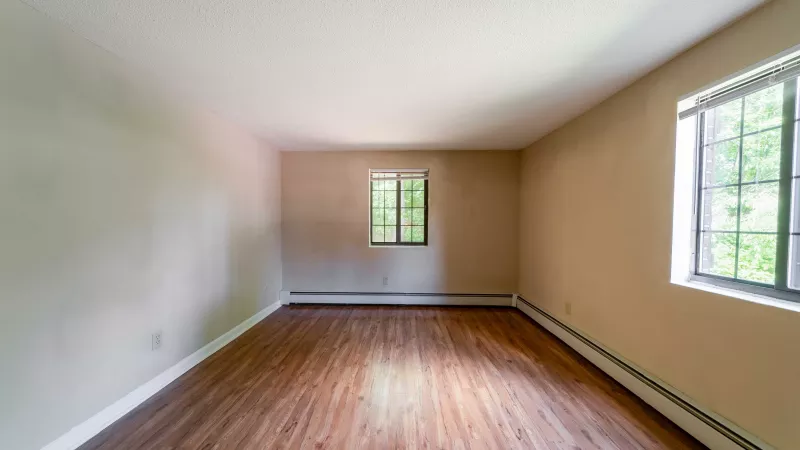 Empty room with warm tan walls, wood-grain floor, and two windows showing green trees.