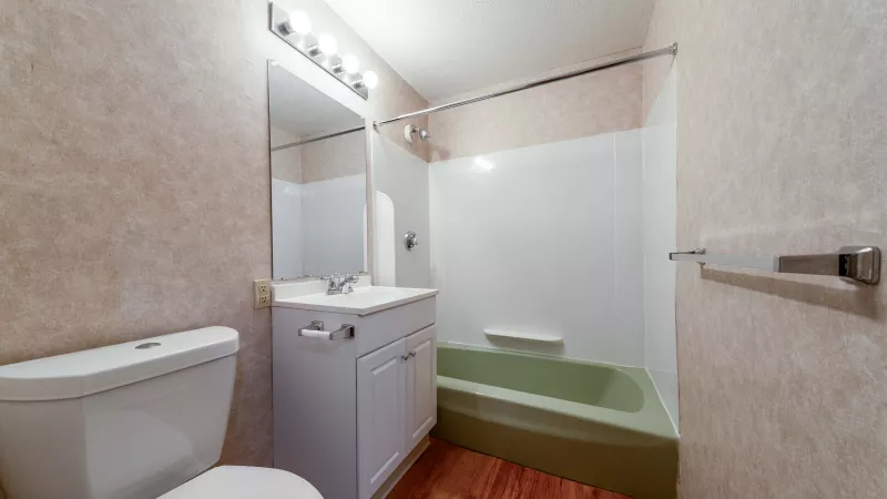 Small bathroom with a white toilet, white vanity, mirror, and an olive green bathtub, all on wood-look flooring.
