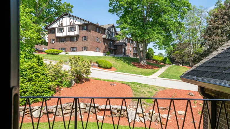 Balcony view of a large, Tudor-style brick building, surrounded by green lawns, trees, and red mulch under a clear sky.