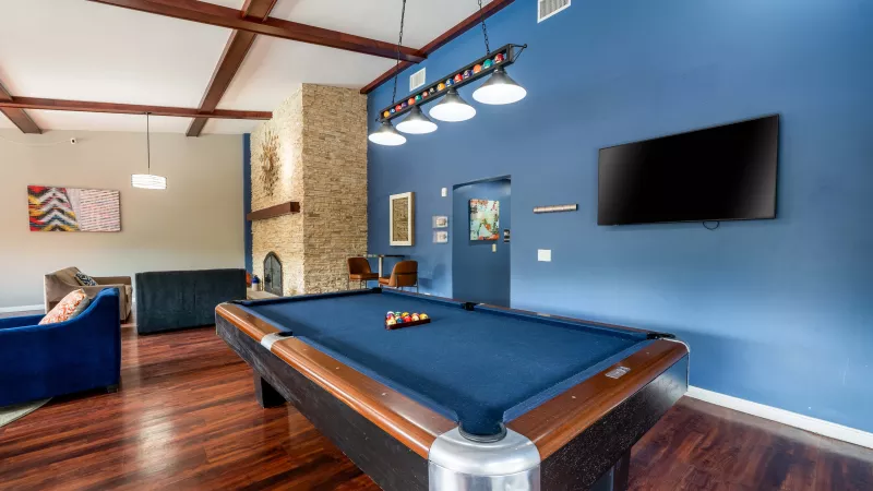 Recreational room with a blue felt pool table, hardwood floors, exposed ceiling beams, a stone fireplace, and a blue accent wall with a TV.