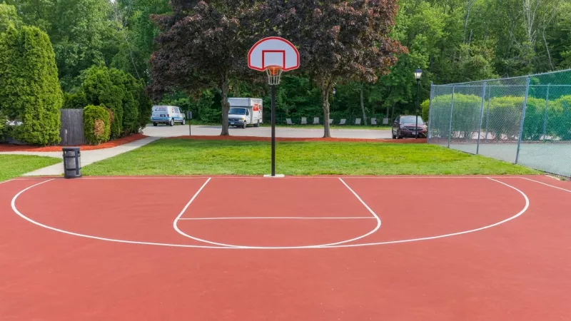 Empty outdoor basketball court with a red surface, white lines, and a single hoop, surrounded by green trees.