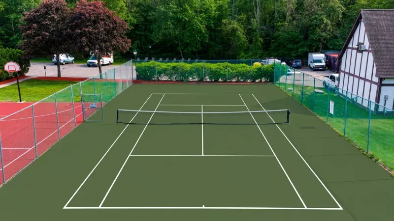 Green tennis court with white lines and net, fenced, with a basketball court and building in the background.
