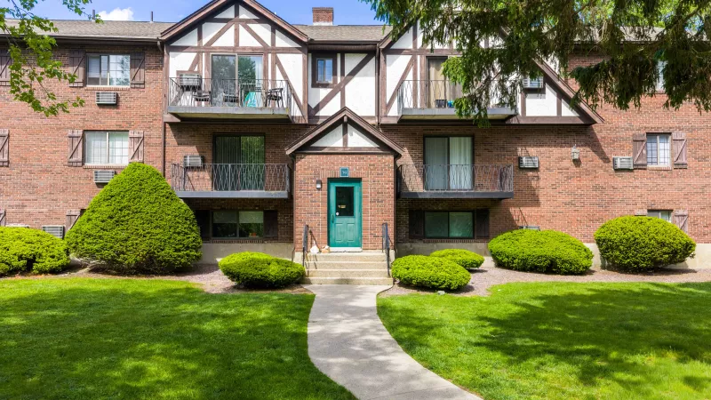 Apartment building with brick facade, Tudor-style gables, a teal entrance, and well-maintained green landscaping.