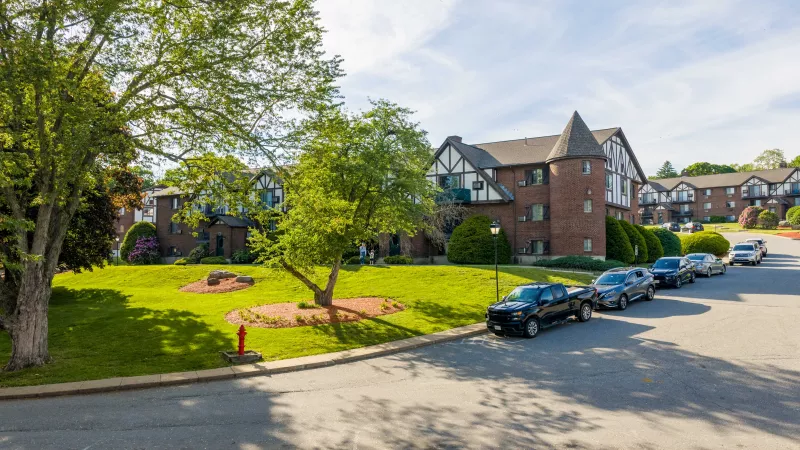 Photograph: Sunny residential street with brick apartment buildings, one featuring a turret, green lawns, trees, and parked cars.