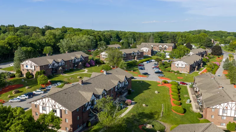 Aerial photo of an apartment community with red brick buildings, white trim, green lawns, and parked cars under a blue sky.