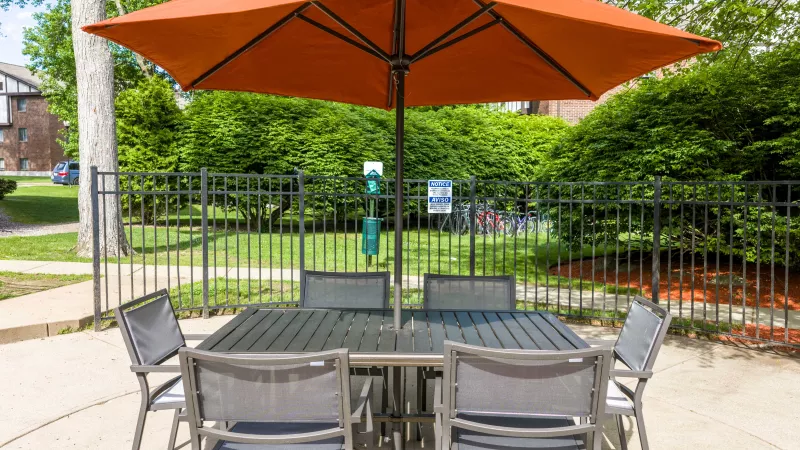 Photograph of an outdoor patio with dark gray table, six chairs, and a large orange umbrella by a fence and landscaping.