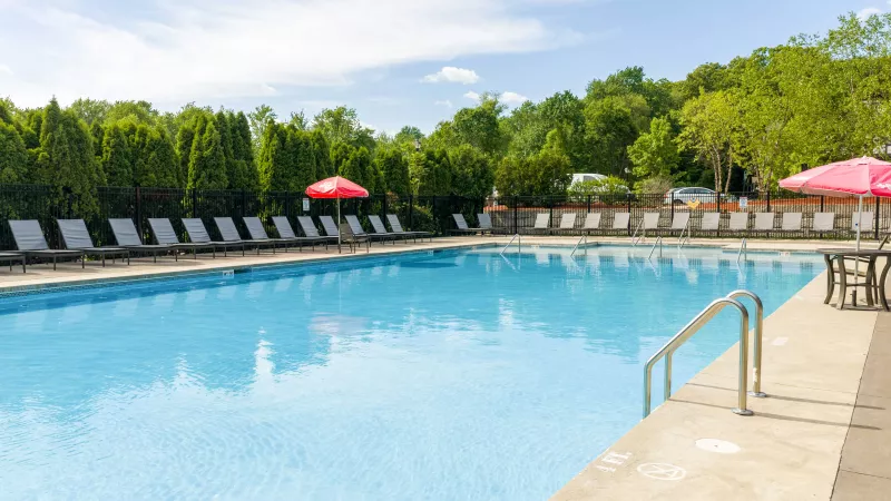 Outdoor swimming pool with clear blue water, surrounded by lounge chairs, red umbrellas, and lush green trees.