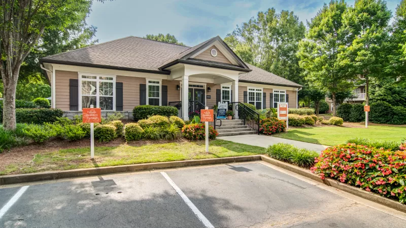 Photograph of a tan building with a dark roof and porch, surrounded by green trees, shrubs, and a paved parking lot.