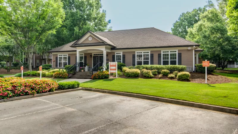 Single-story tan building with dark roof, green landscaping, and red flowers. Paved parking lot in front.