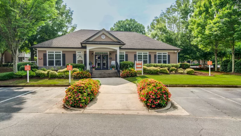 Clubhouse or office building with dark siding, white trim, gabled entrance, green trees, and colorful shrubs.