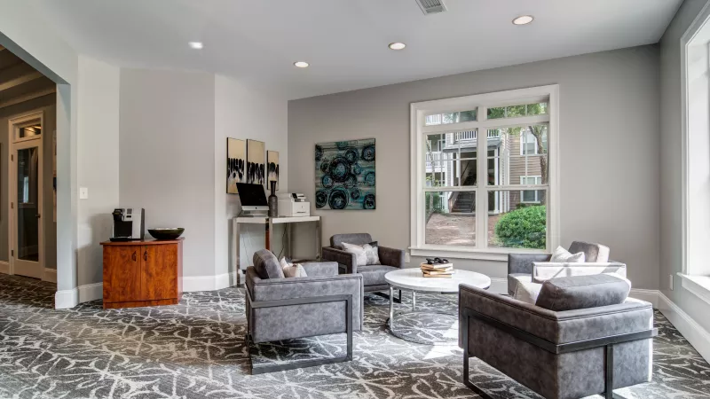 Bright, modern lounge with grey armchairs, coffee table, patterned carpet, computer station, and large window.