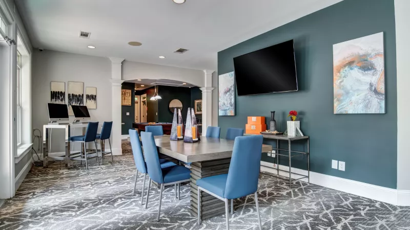 Modern common area with a large table, blue chairs, TV on a teal wall, computer workstations, and a patterned rug.