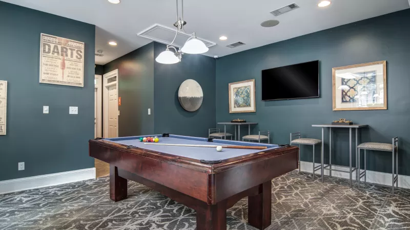 Photograph of a stylish recreation room with a pool table, dark teal walls, patterned rug, TV, and high-top tables.