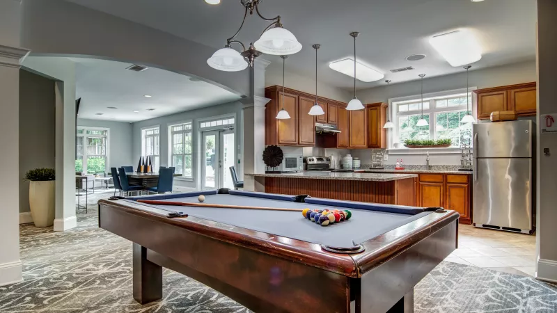 Clubhouse common room featuring a pool table, full kitchen with wood cabinets, and a sunlit dining area beyond an archway.