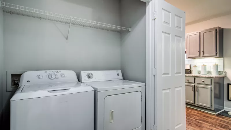 White washer and dryer in a grey laundry closet with a wire shelf. A kitchen with grey cabinets is visible through an open door.