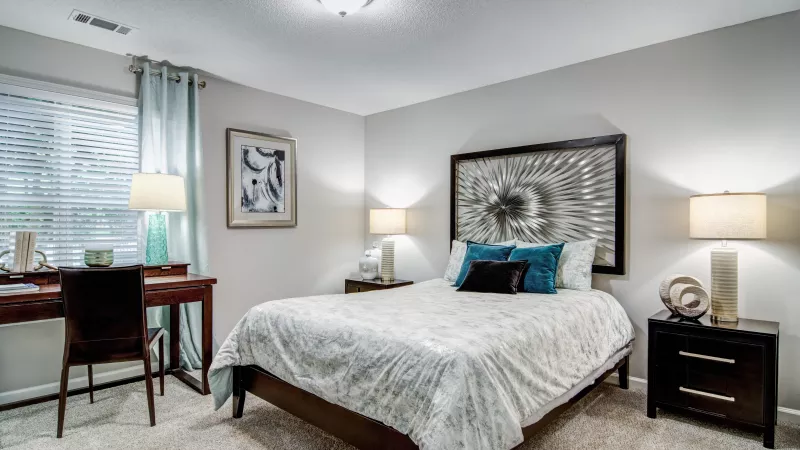 Well-furnished bedroom with a bed, two nightstands, lamps, abstract art, and a desk by a window with teal curtains.