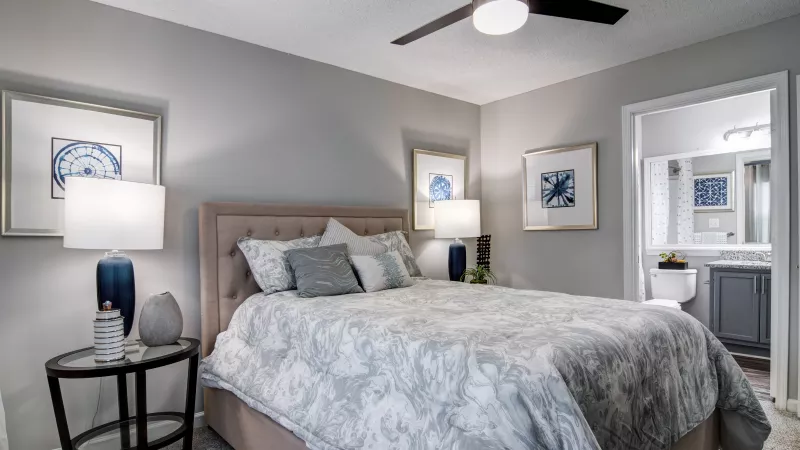 Modern bedroom with a beige bed, blue lamps, framed art, and an open doorway leading to a bathroom.