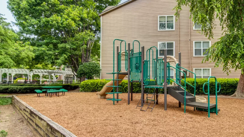 Empty playground with green and gray equipment on wood chips, flanked by an apartment building and trees. Photograph.
